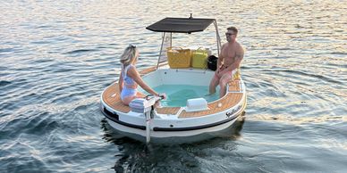 A couple enjoys a small round boat with a built-in hot tub on calm water.