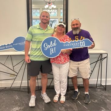 Three people smiling and holding a "Sold It!" sign in front of a mirror indoors.