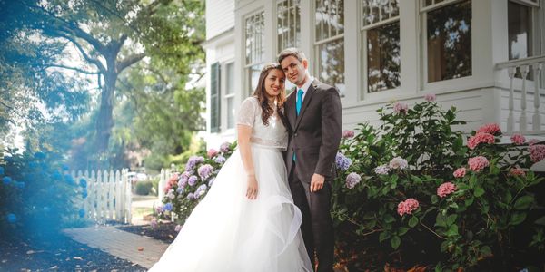 Newlywed couple posing in wedding attire beside a white house with blooming flowers.