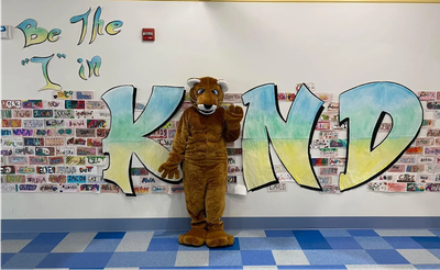School mascot standing in front of a colorful 'Be The I in KIND' display.