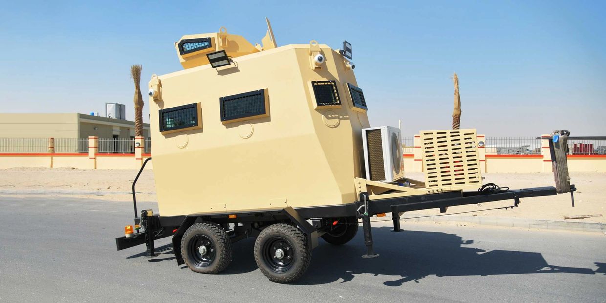 A beige armored trailer with black wheels parked on a road in a desert environment.
