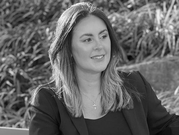 Woman in black dress and blazer sitting on a park bench in sunlight.