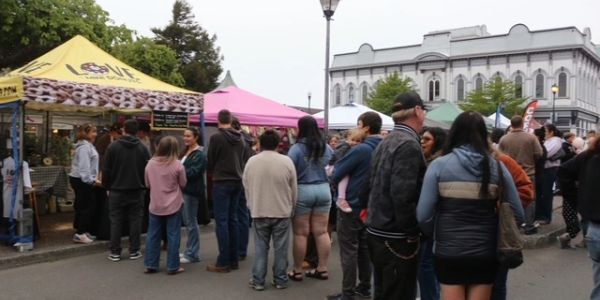 People queue at outdoor market stalls on a cloudy day.
