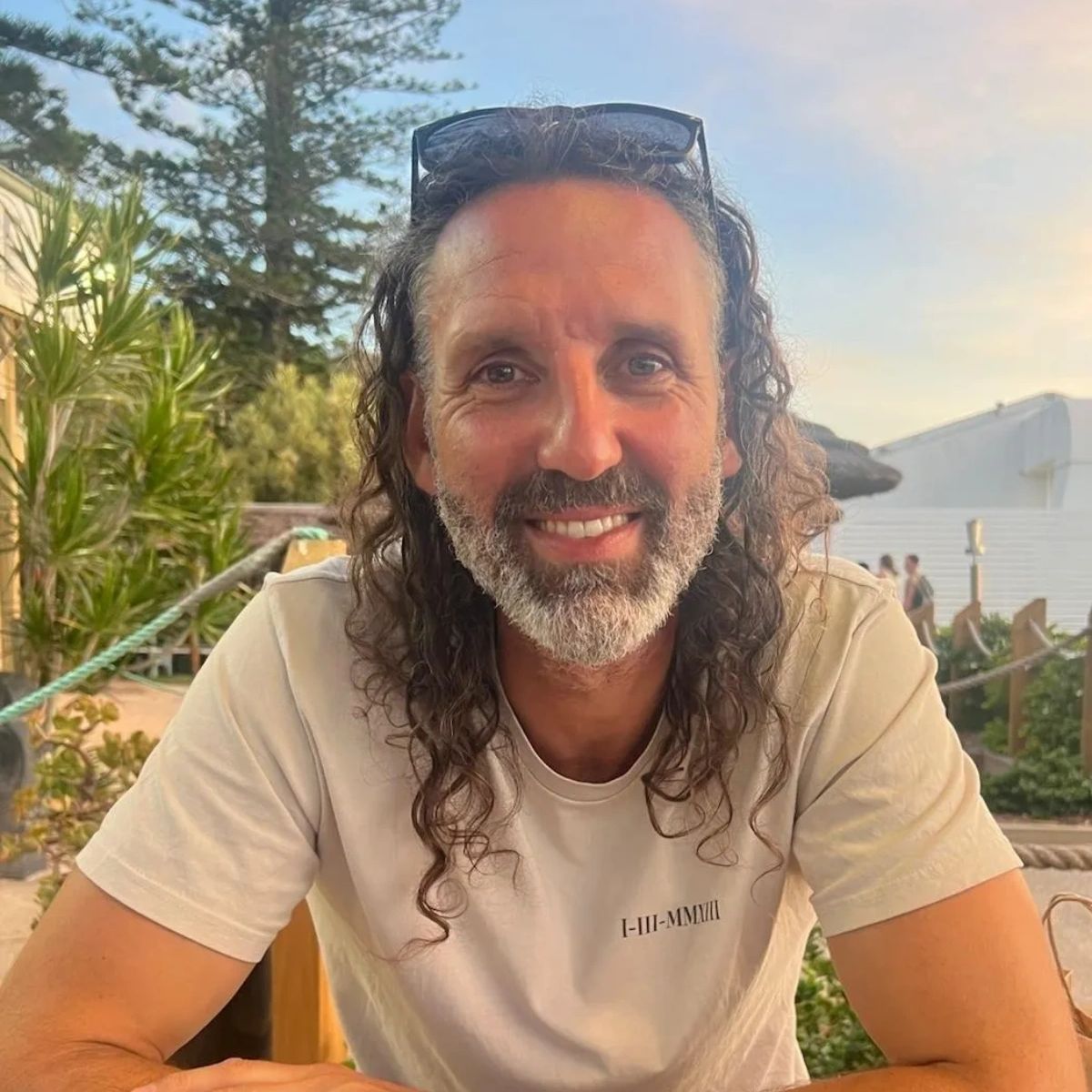 Smiling man with curly hair and beard outdoors at sunset.