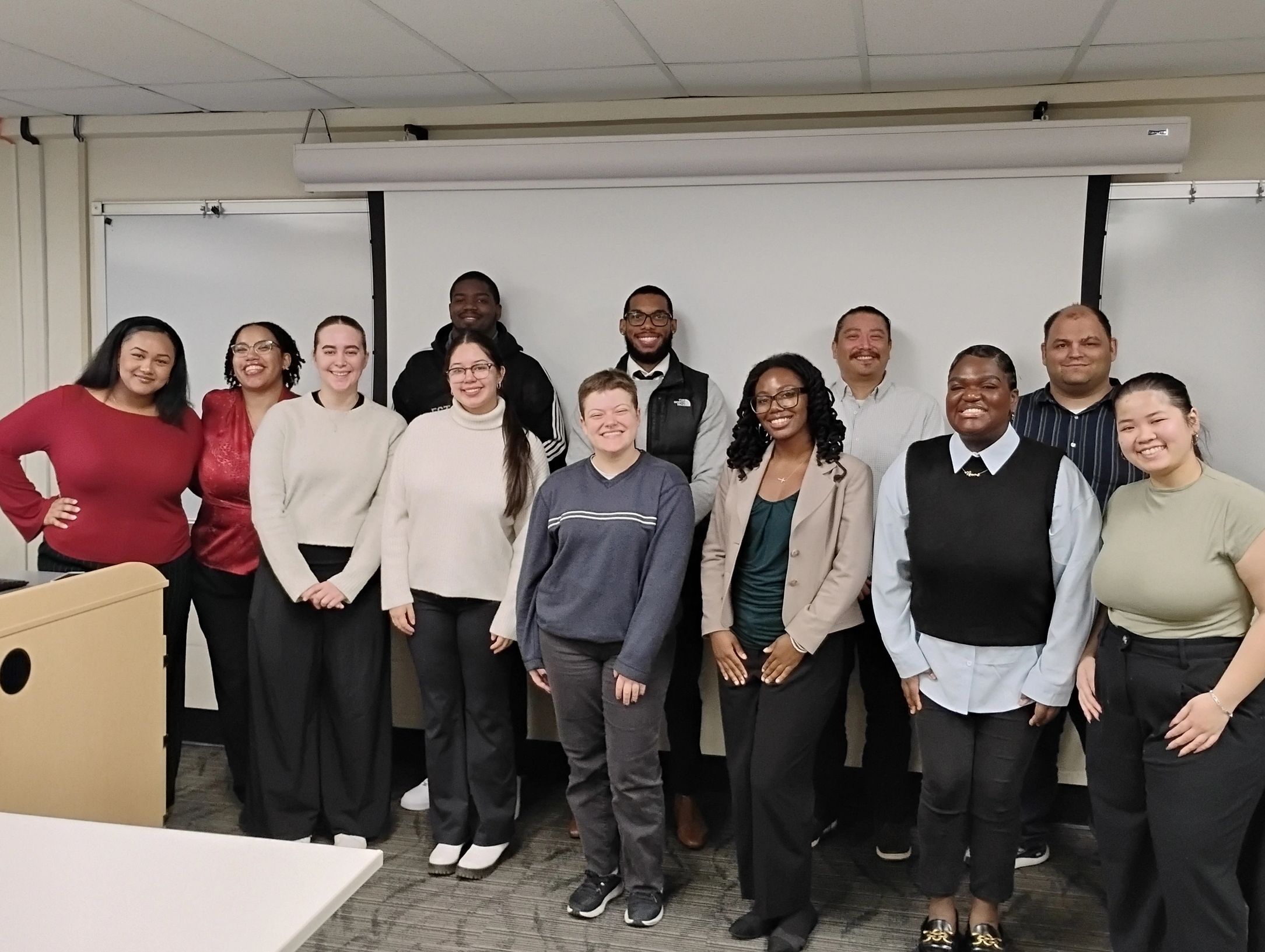 Group of smiling people standing in front of a whiteboard in a classroom.