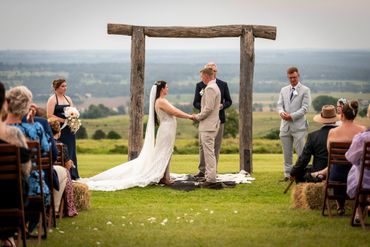 Outdoor wedding ceremony with bride and groom holding hands under a rustic wooden arch.