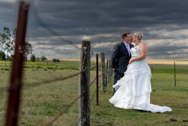 Newlyweds sharing a kiss in a rustic field under a dramatic cloudy sky.