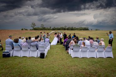 Outdoor wedding ceremony under dramatic cloudy sky with guests seated and couple exchanging vows.