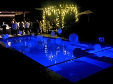 White helium balloons in pool with blue lights and fairy lights on gazebo.