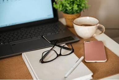 Workspace with laptop, glasses, notebook, pen, phone, and coffee.
