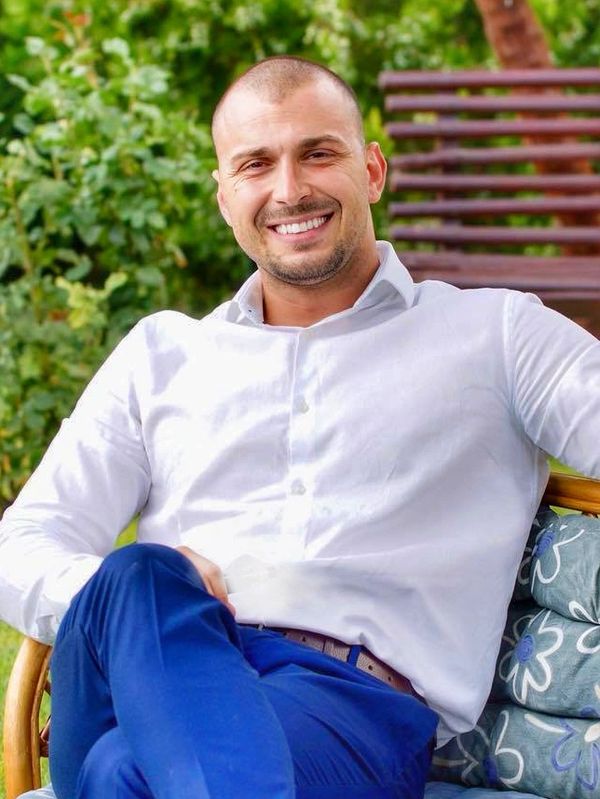 Smiling man in white shirt and blue pants sitting outdoors on a chair.