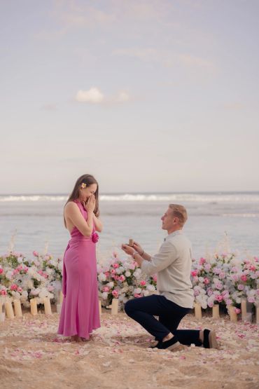 Man proposes to woman on beach surrounded by flowers.