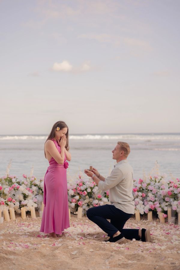 Man proposes to woman on beach surrounded by flowers.