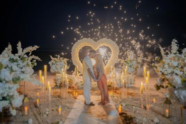 Couple kissing at romantic beach setup with heart and fireworks.