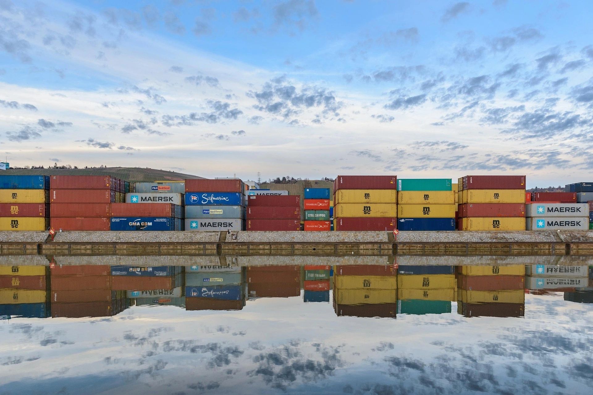 Shipping containers stacked by the water with a reflection on a calm day.