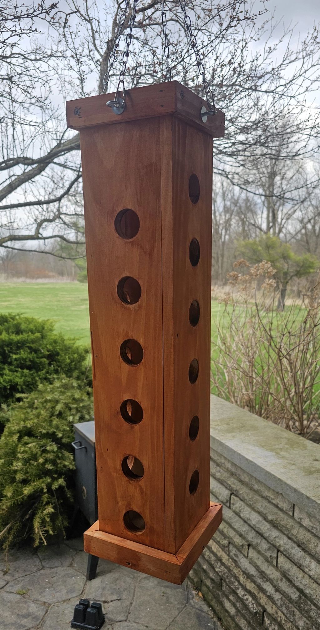 Tall wooden bird feeder with multiple round openings hanging outdoors.