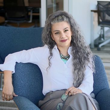 A woman with long gray hair sitting on a blue chair, smiling.