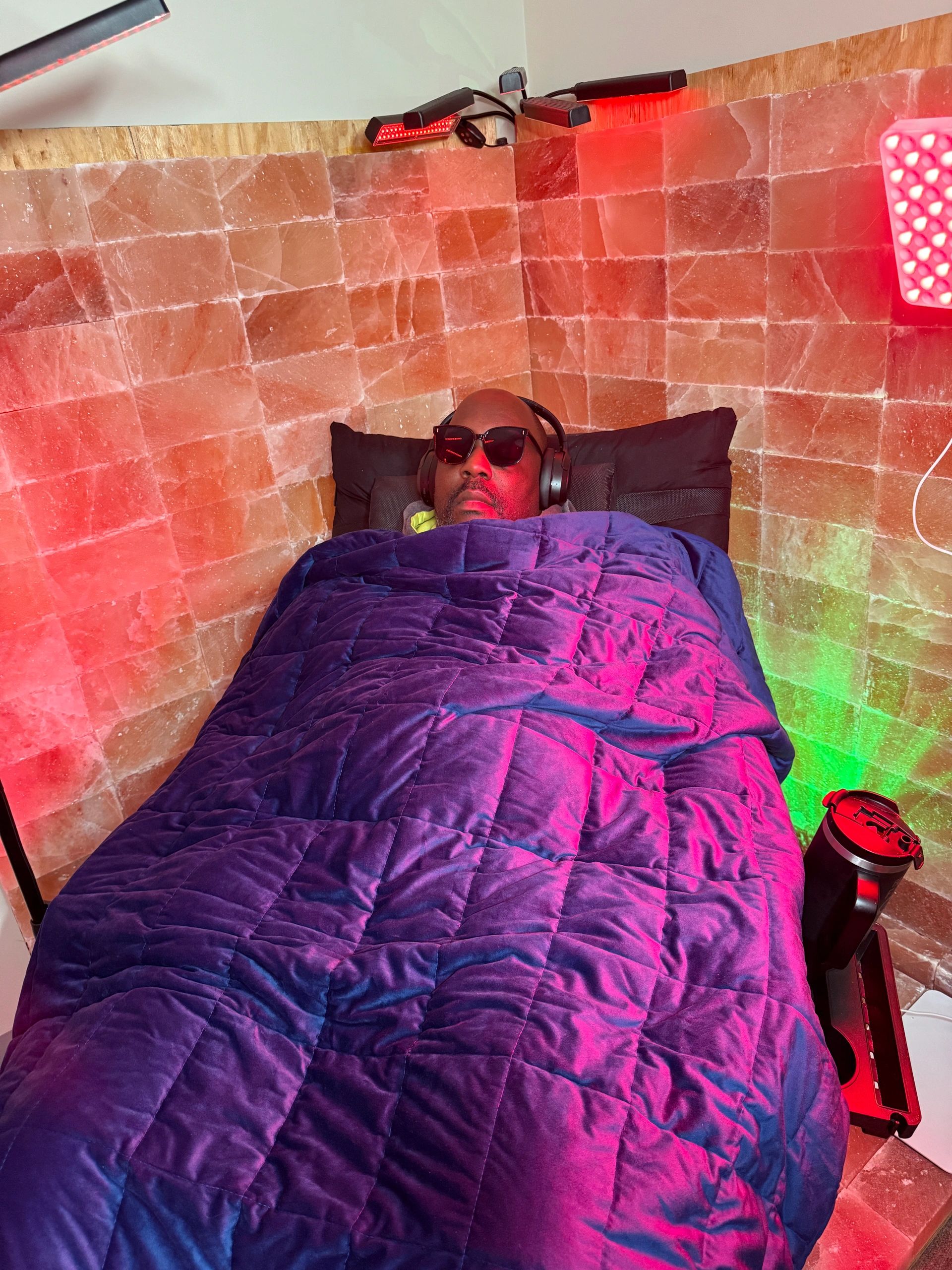 A person relaxing under a blue blanket with sunglasses and headphones in a room with red lighting.