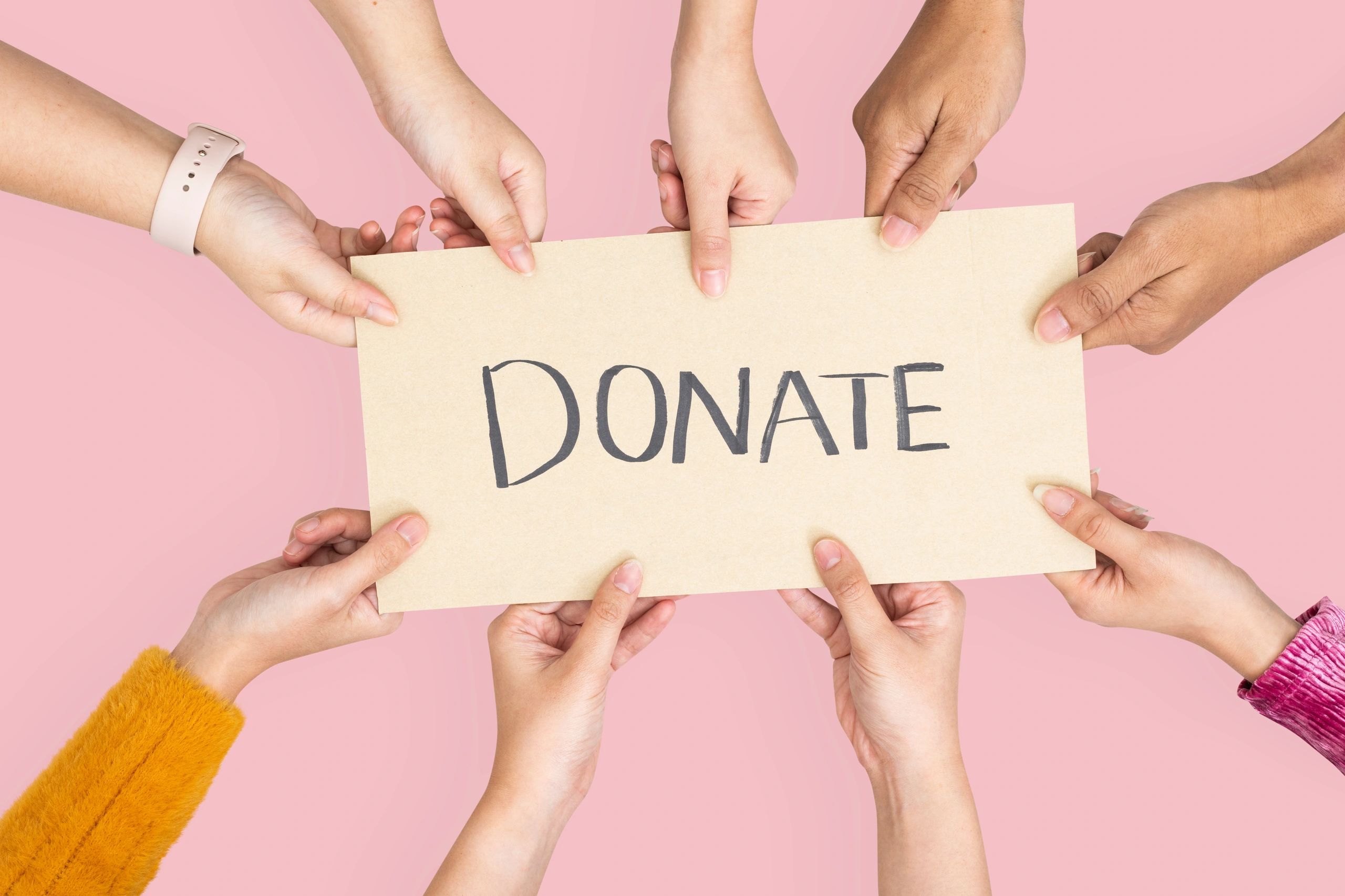 Multiple hands holding a sign that says 'DONATE' on a pink background.