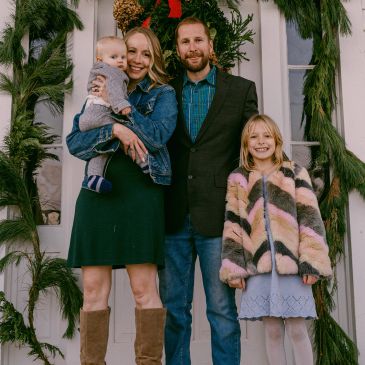A smiling family of four posing in front of a decorated doorway with greenery and a red bow.