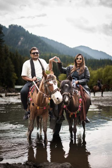 Couple on horseback making a heart with hands in a scenic mountain river.