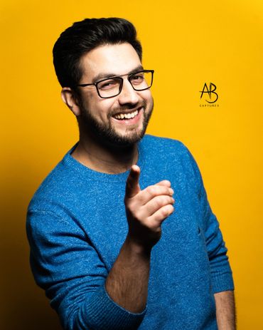 Smiling man in blue sweater pointing forward against a yellow background.