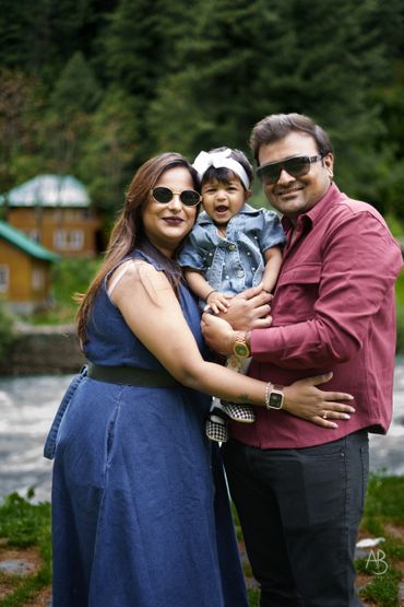 A smiling family of three posing outdoors with greenery in the background.