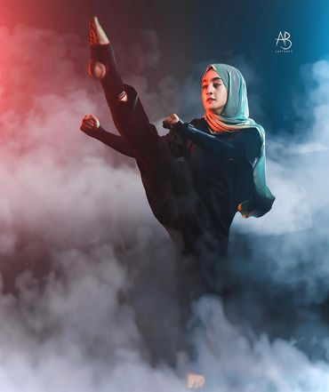 A woman in a hijab performs a high martial arts kick amidst fog and colorful lighting.