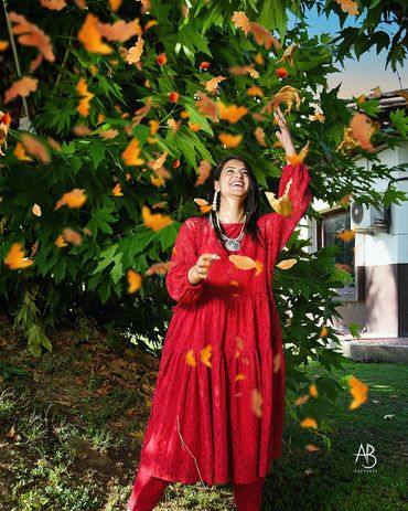 A joyful woman in a red dress throws autumn leaves under a green tree.