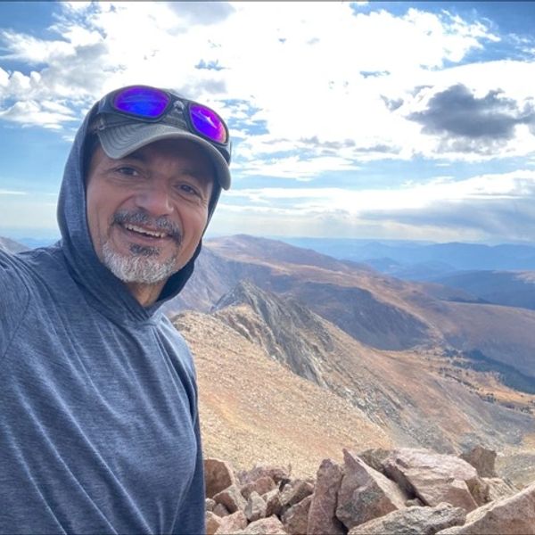 Man in hoodie and cap smiling with mountain landscape in background.
