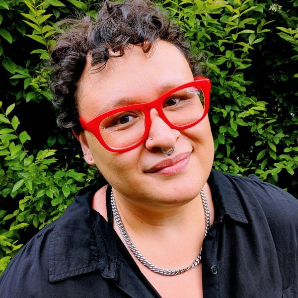 Person with curly hair, red glasses, and a silver chain necklace smiling outdoors.