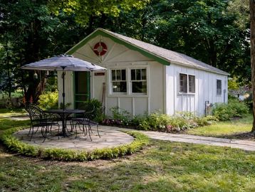 Cozy white garden shed with outdoor seating and umbrella in lush greenery.
