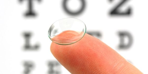 A soft contact lens resting on a fingertip with an eye chart in the background.
