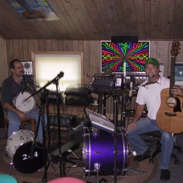 Two musicians with instruments in a cozy wooden room filled with music equipment.