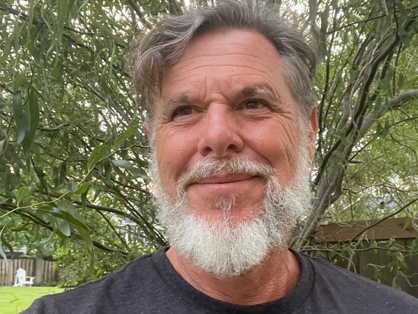 Smiling man with white beard outdoors near trees.