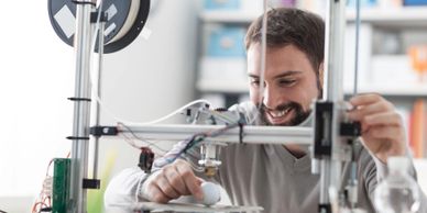 Man smiling while working on a 3D printer in a bright room.