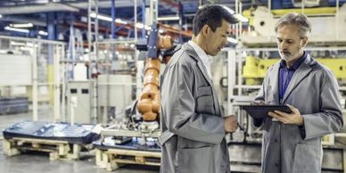 Two engineers discussing data on a tablet in a factory with robotic machinery.