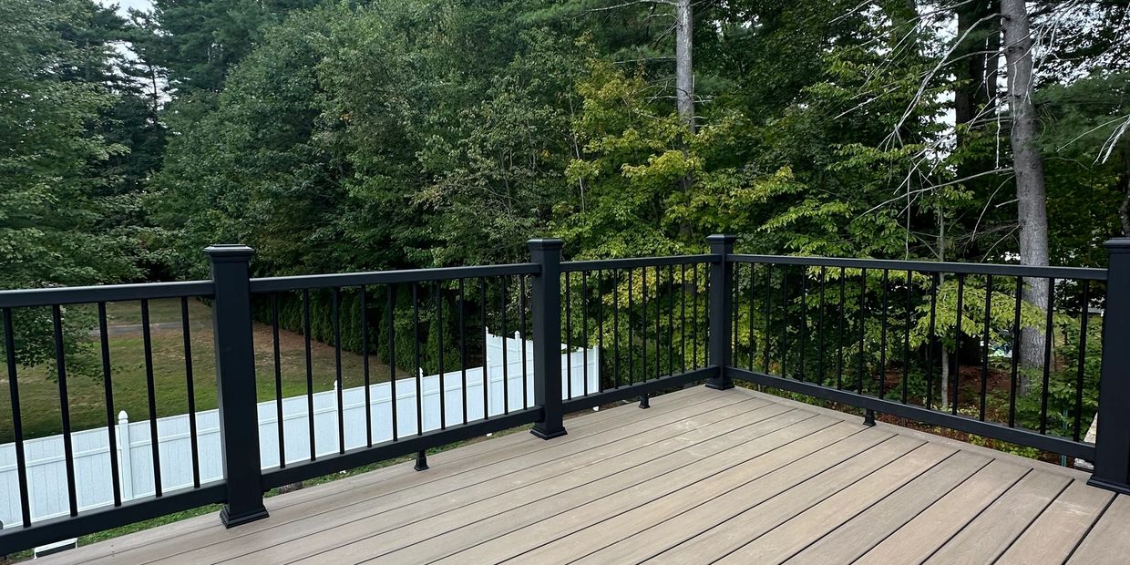 Empty deck with black railing overlooking green trees and white fence.