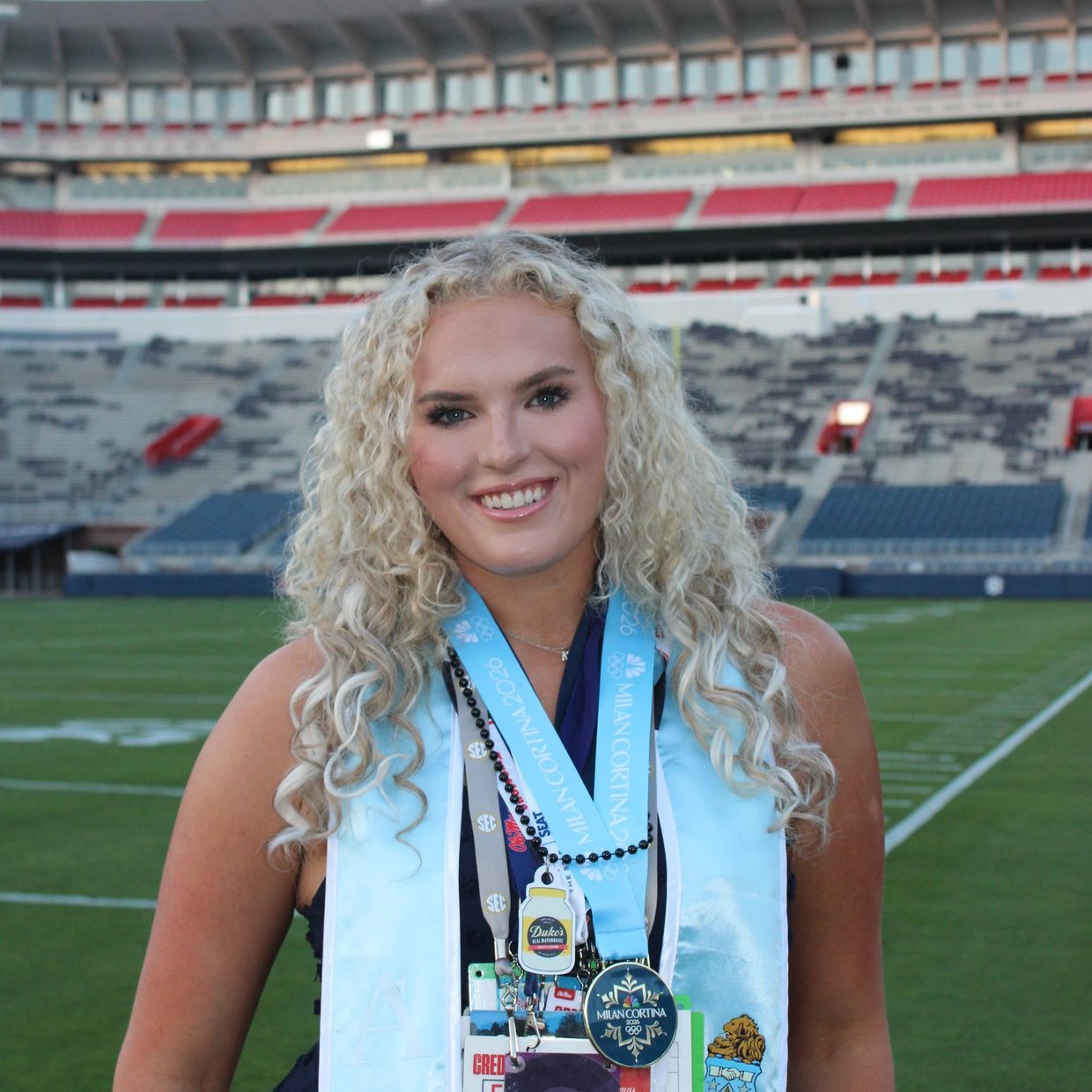 Smiling graduate with medals and lanyards at a sports stadium.