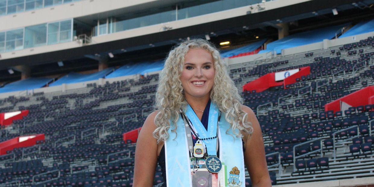 A smiling graduate with curly blonde hair stands in a large stadium.