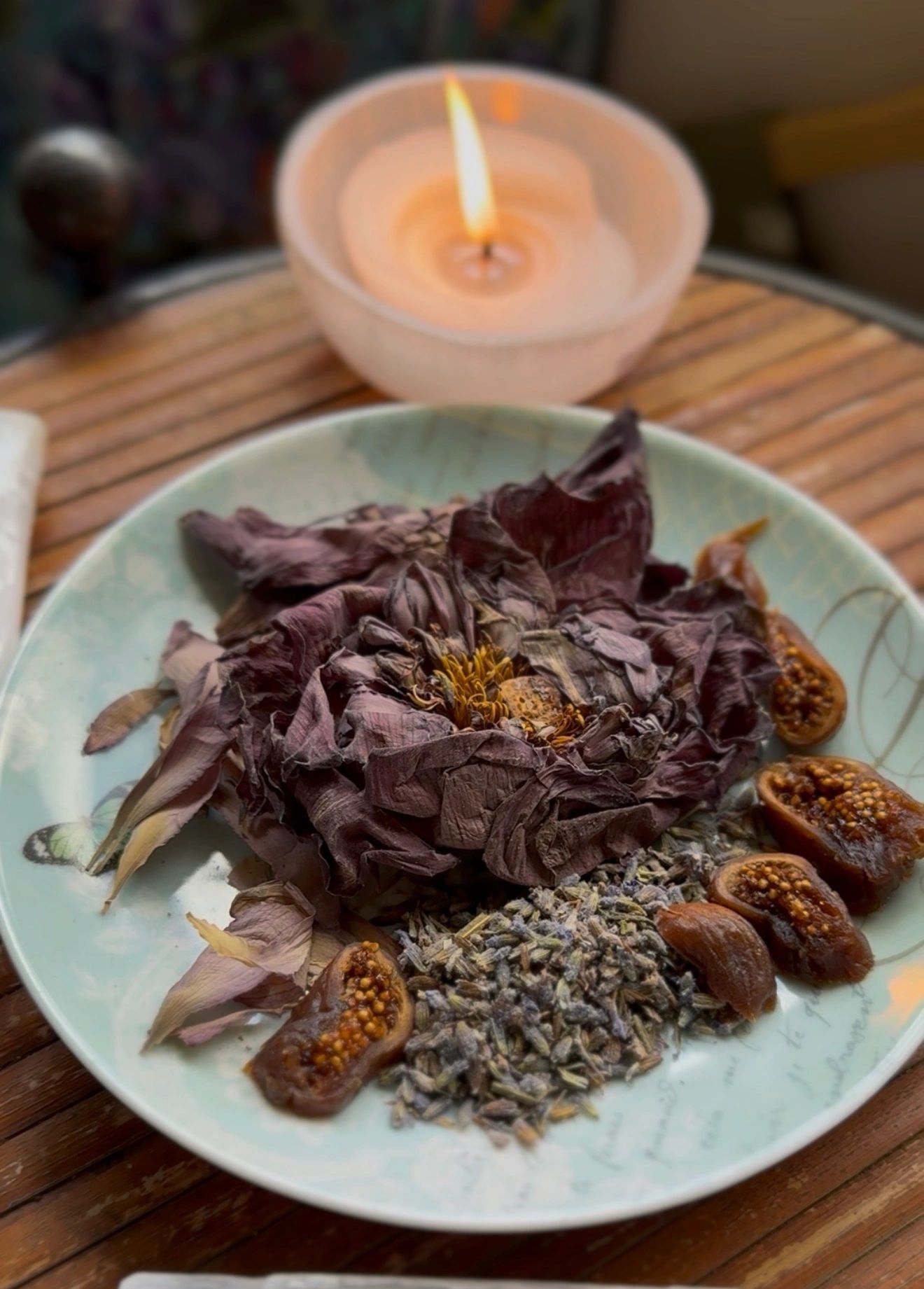 Dried flower petals, herbs, and fig slices on a plate with a lit candle in the background.