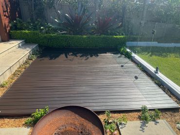 A wooden deck in a backyard with plants and glass fencing.