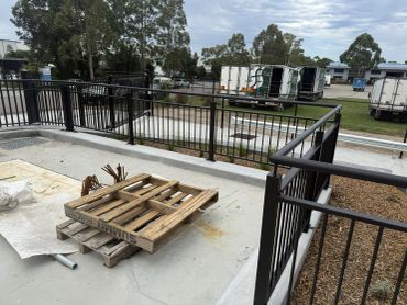 Metal railings around a concrete area with wooden pallets and trucks in the background.