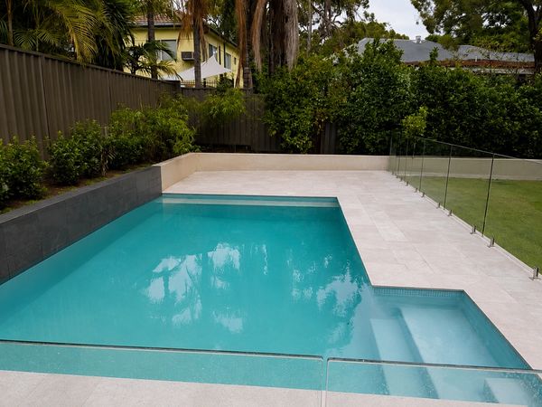 Modern backyard swimming pool with glass fence and greenery.
