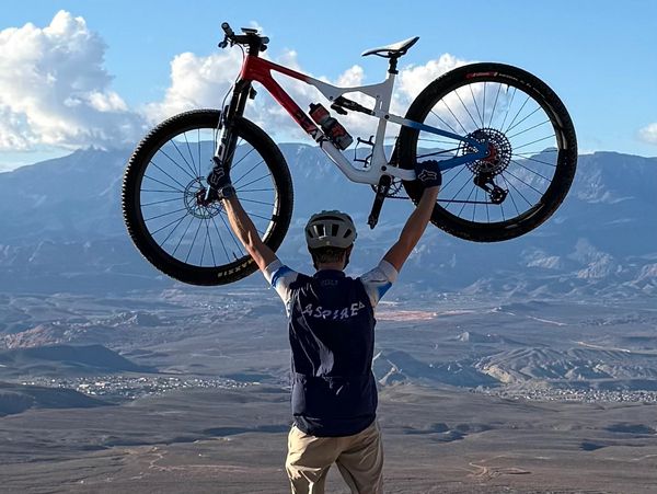 A cyclist lifts his bike triumphantly against a mountain backdrop.