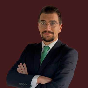 Confident man in a suit with glasses and a green tie against a maroon background.