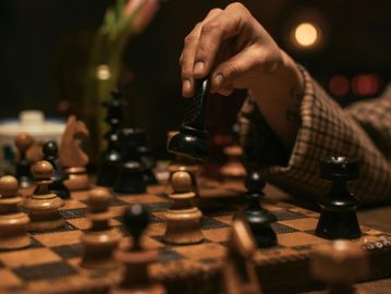 Hand moving a black chess piece on a wooden chessboard in dim lighting.