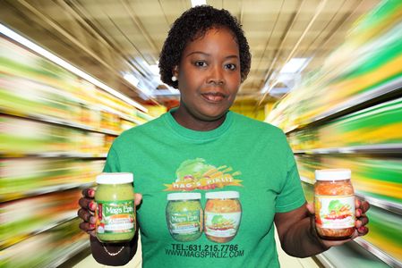 A person wearing a green shirt and holding a bottle of Mag's Pikliz and a bottle of Mag's Epis