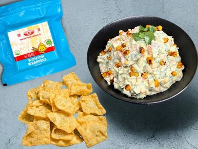 Bowl of creamy dip with roasted corn and chips beside wonton wrappers packet.