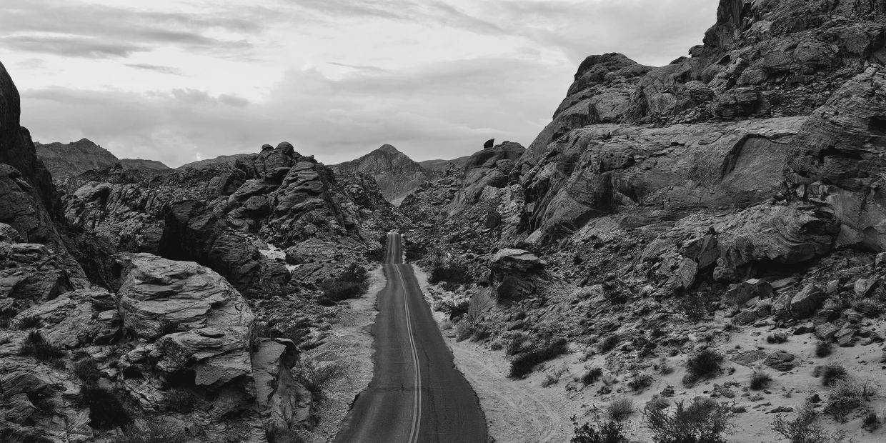 Desert road cutting through rocky mountains under a cloudy sky in black and white.